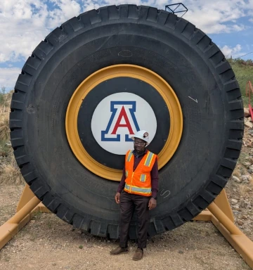 A man standing in front of a giant tire with U of A Logo. The man is wearing a vest and a hard hat.